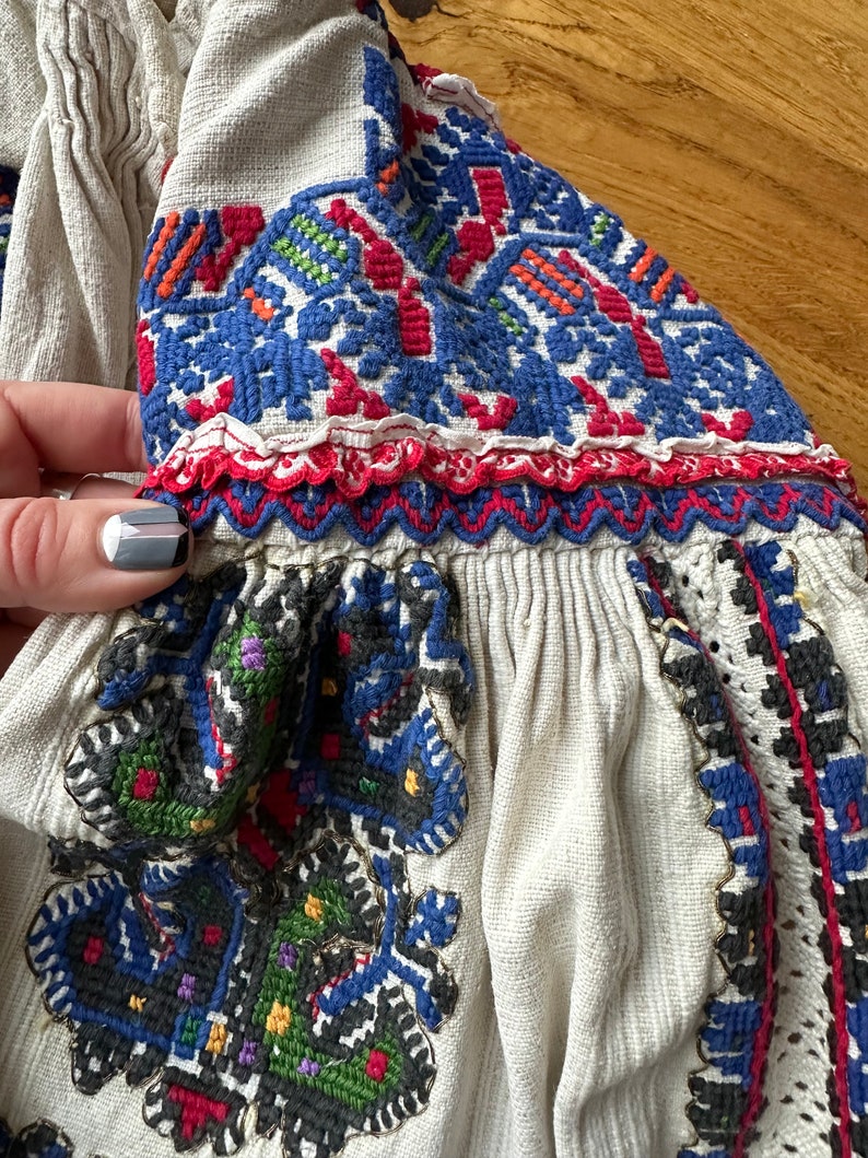 May include: A white linen blouse with colorful embroidered details. The embroidery features geometric patterns in shades of blue, red, green, and yellow.