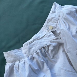 May include: Close-up of a white cotton shirt collar and shoulder. The shirt has a button-down collar and a button closure at the neck. The fabric is slightly wrinkled, suggesting it is made of a soft material.