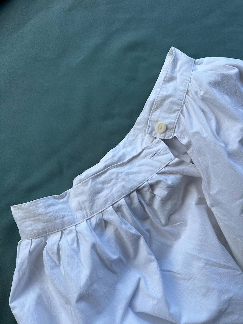 May include: Close-up of a white cotton shirt collar and shoulder. The shirt has a button-down collar and a button closure at the neck. The fabric is slightly wrinkled, suggesting it is made of a soft material.