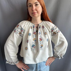 May include: A white embroidered blouse with a floral pattern in shades of blue, red, yellow, and green. The blouse has long sleeves and a gathered neckline.