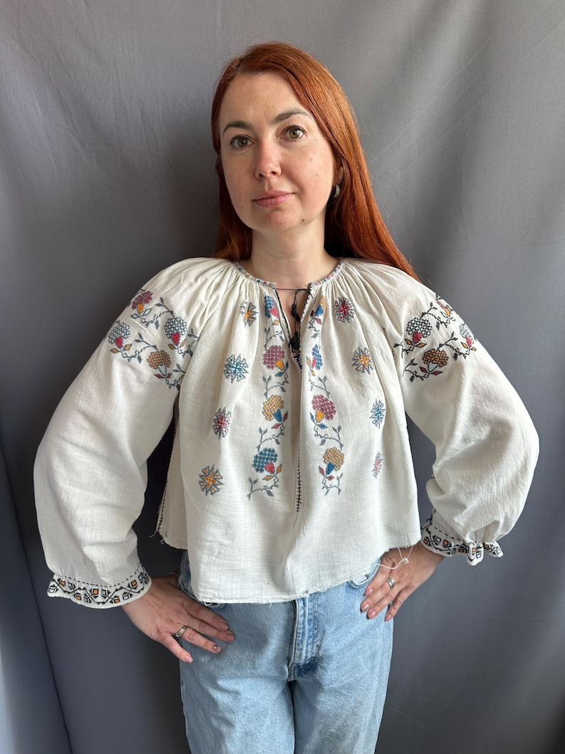 May include: A white embroidered blouse with a floral pattern in shades of blue, red, yellow, and green. The blouse has long sleeves and a gathered neckline.