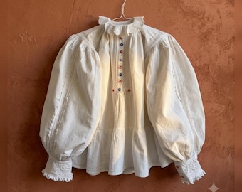 May include: A white long-sleeve blouse featuring a high ruffled collar, voluminous puff sleeves, and intricate lace detailing along the shoulders and cuffs. The front placket is adorned with a vertical row of small, multicolored circular buttons.