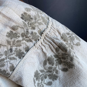 May include: A light beige linen garment featuring intricate grey cross-stitch floral embroidery. The fabric displays a visible woven texture, with gathered detailing along the seam accented by a decorative braided trim running horizontally.