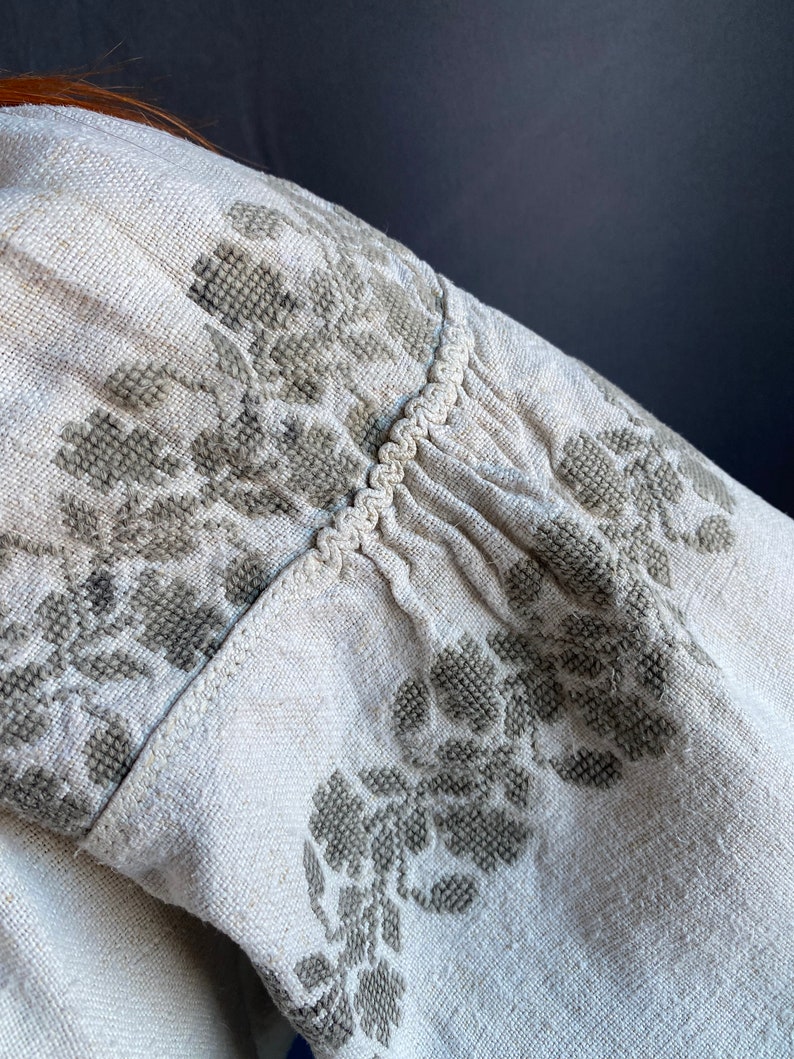 May include: A light beige linen garment featuring intricate grey cross-stitch floral embroidery. The fabric displays a visible woven texture, with gathered detailing along the seam accented by a decorative braided trim running horizontally.
