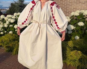 May include: A cream-colored midi dress featuring voluminous long sleeves with intricate red and blue geometric embroidery. The garment has a gathered neckline with a thin tie closure and a simple waist tie, showcasing a loose, flowing silhouette.