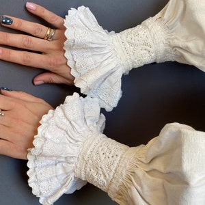 K&ouml;nnte beinhalten: Wei&szlig;e Baumwollbluse mit langen &Auml;rmeln und Spitzenbesatz an den Manschetten. Die Manschetten haben einen gerafften Look und sind mit filigranen Stickereien verziert.