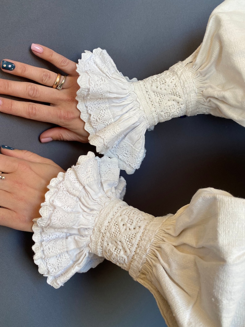 K&ouml;nnte beinhalten: Wei&szlig;e Baumwollbluse mit langen &Auml;rmeln und Spitzenbesatz an den Manschetten. Die Manschetten haben einen gerafften Look und sind mit filigranen Stickereien verziert.