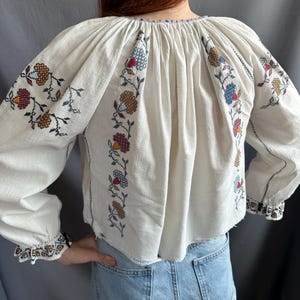 May include: White embroidered blouse with a gathered neckline and long sleeves. The embroidery features floral designs in shades of red, blue, orange, and brown.