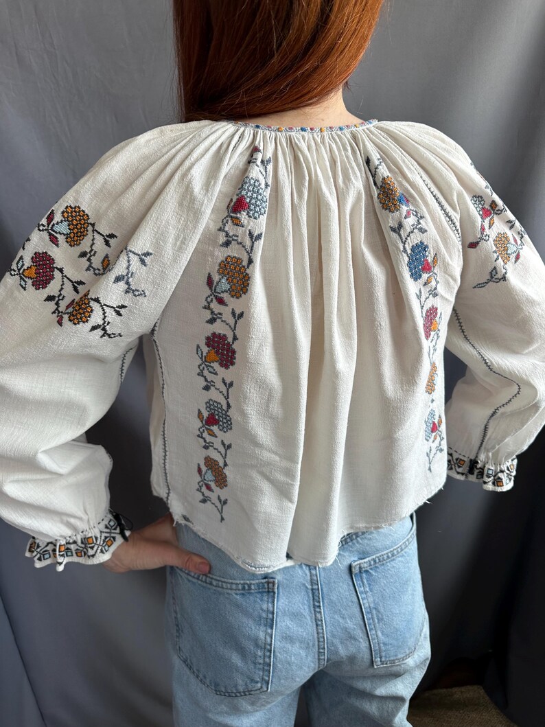 May include: White embroidered blouse with a gathered neckline and long sleeves. The embroidery features floral designs in shades of red, blue, orange, and brown.