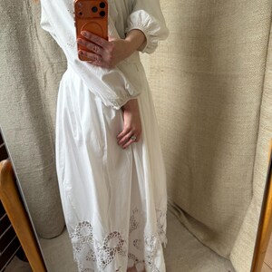 May include: White long-sleeve dress with intricate eyelet embroidery on the bodice and sleeves. The dress features a full skirt with a scalloped, lace-like hem. The dress has a high neckline and a relaxed fit, suitable for various occasions.