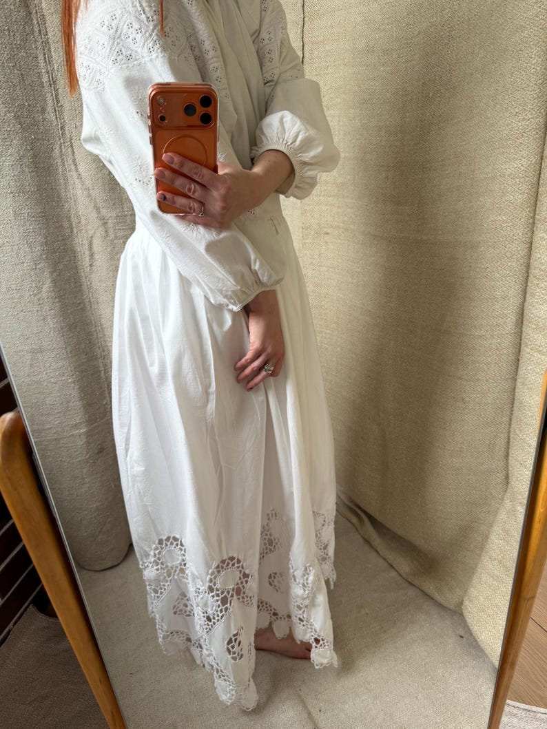 May include: White long-sleeve dress with intricate eyelet embroidery on the bodice and sleeves. The dress features a full skirt with a scalloped, lace-like hem. The dress has a high neckline and a relaxed fit, suitable for various occasions.