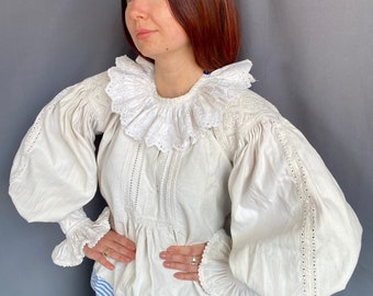 May include: A white linen blouse with a large, ruffled collar and long, puffed sleeves. The blouse has delicate lace detailing throughout.