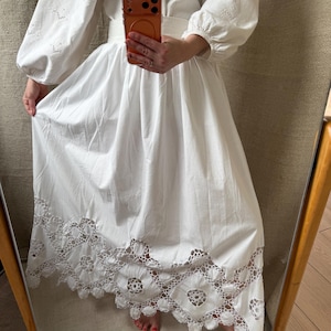 May include: A white dress with long, puffy sleeves and a wide skirt. The sleeves feature intricate cut-out detailing, and the skirt's hem is adorned with a delicate, openwork lace design. The dress is belted at the waist.
