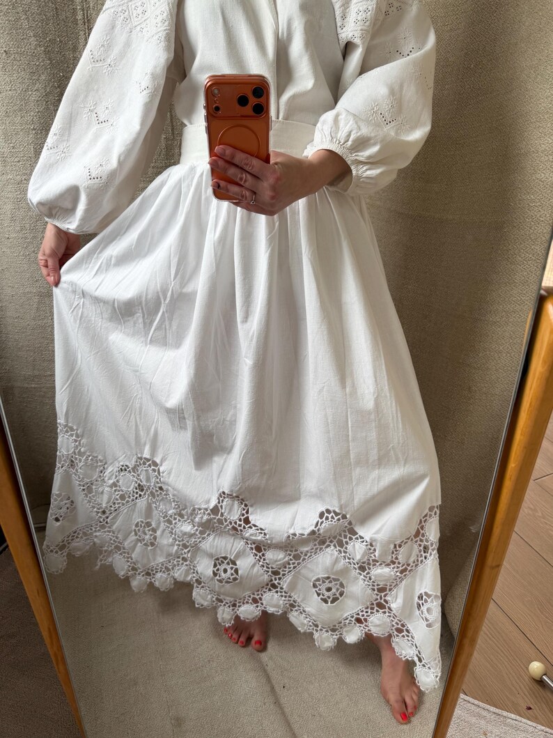 May include: A white dress with long, puffy sleeves and a wide skirt. The sleeves feature intricate cut-out detailing, and the skirt's hem is adorned with a delicate, openwork lace design. The dress is belted at the waist.