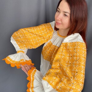 May include: A white blouse with long sleeves and a yellow embroidered pattern. The sleeves have white lace trim and the embroidery features a geometric design.