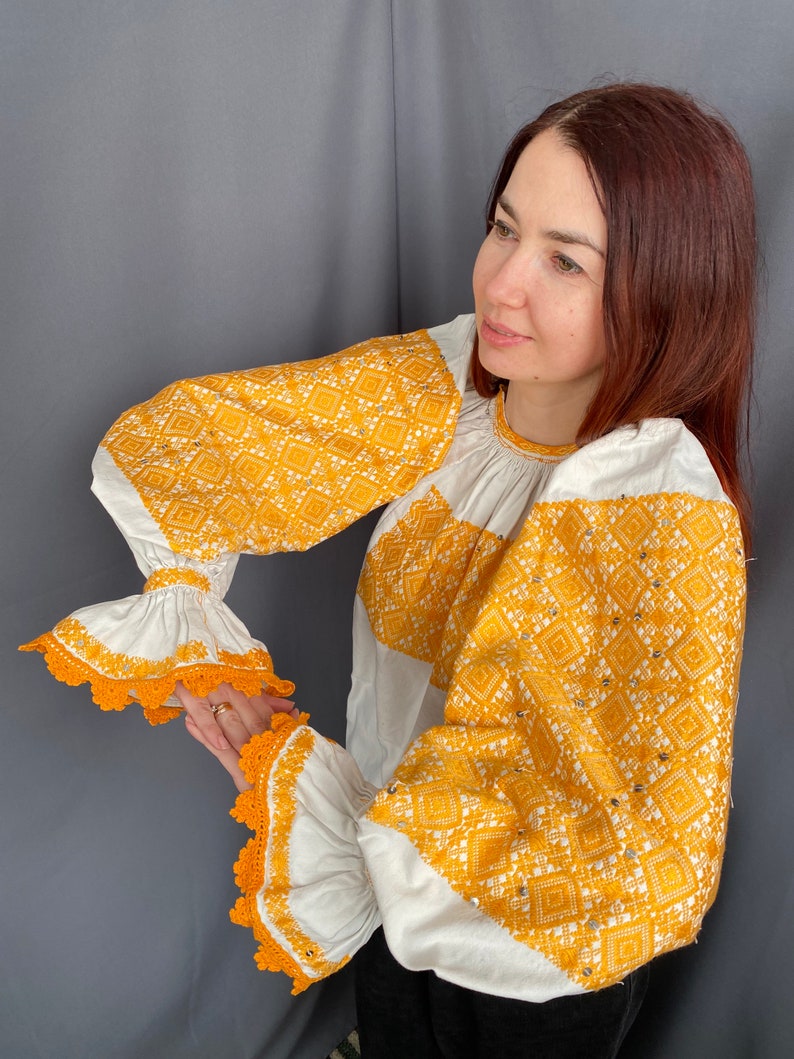 May include: A white blouse with long sleeves and a yellow embroidered pattern. The sleeves have white lace trim and the embroidery features a geometric design.