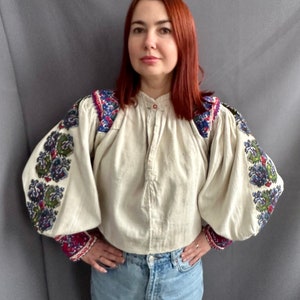 May include: A white linen blouse with embroidered floral patterns in shades of blue, green, red, and purple. The blouse has a button-up front and long, puffy sleeves.