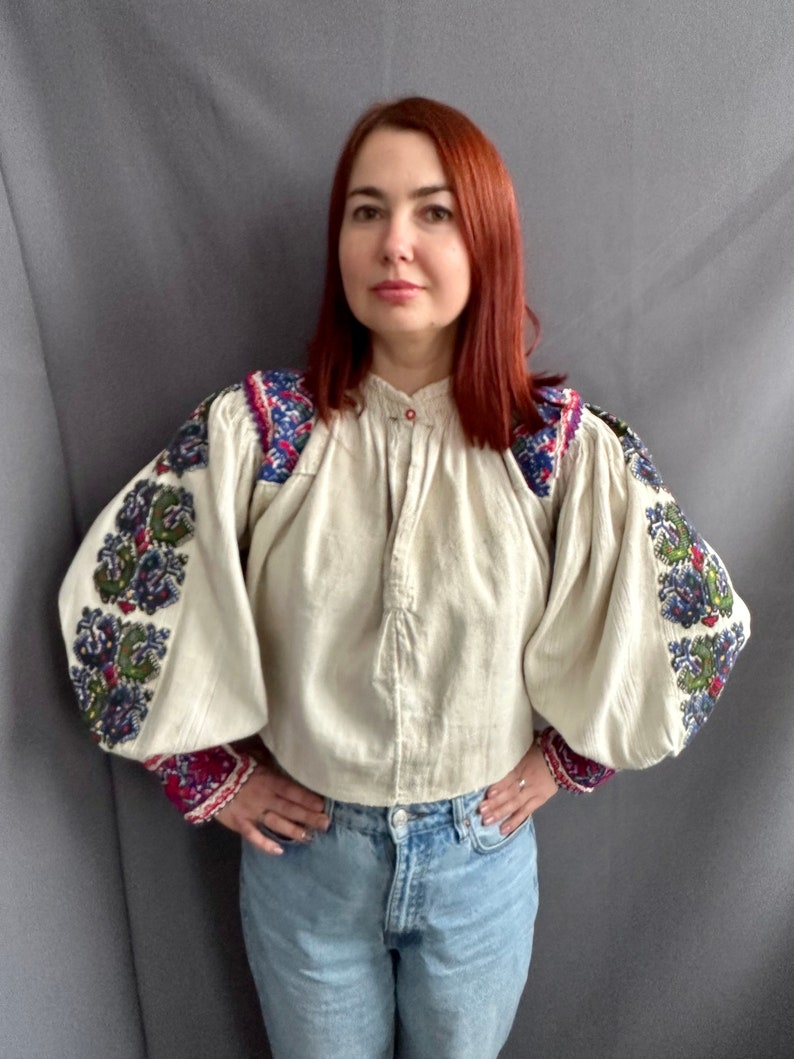 May include: A white linen blouse with embroidered floral patterns in shades of blue, green, red, and purple. The blouse has a button-up front and long, puffy sleeves.