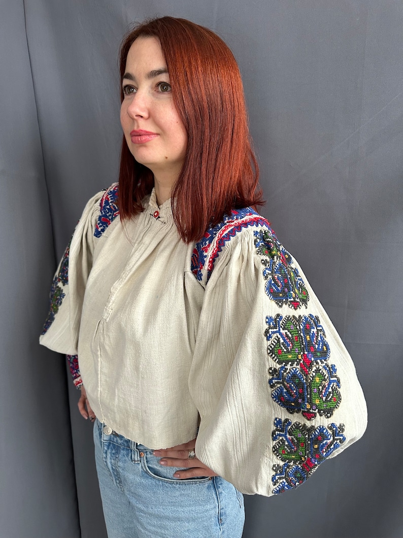 May include: A white linen blouse with embroidered sleeves. The embroidery is in shades of blue, green, red, and black. The blouse has a round neckline and long sleeves.