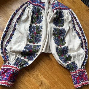 May include: A white embroidered blouse with long sleeves and a button-up collar. The embroidery is in shades of blue, red, green, and black, and features floral patterns. The blouse is made of a lightweight fabric and has a relaxed fit.