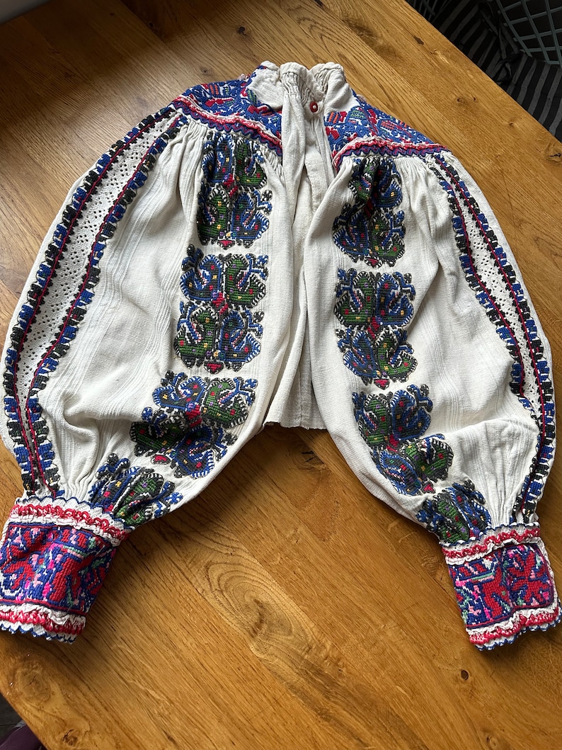 May include: A white embroidered blouse with long sleeves and a button-up collar. The embroidery is in shades of blue, red, green, and black, and features floral patterns. The blouse is made of a lightweight fabric and has a relaxed fit.