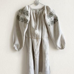 May include: A beige long-sleeved tunic featuring a split neckline and a thin waist tie. The garment is decorated with intricate green floral embroidery across the shoulders and upper sleeves, and it hangs from a metal hanger.