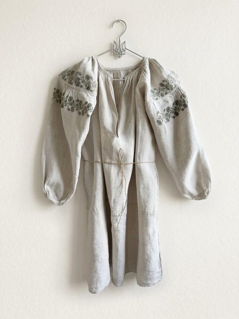 May include: A beige long-sleeved tunic featuring a split neckline and a thin waist tie. The garment is decorated with intricate green floral embroidery across the shoulders and upper sleeves, and it hangs from a metal hanger.