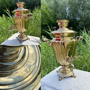 May include: A gold samovar with intricate details and a spout on the side. The samovar is sitting on a white tablecloth with green foliage in the background. The samovar has a round base and a tall, cylindrical body with a domed lid. The inscription "Баташевъ & Ко" is visible on the base.
