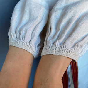 May include: A long-sleeved shirt made of off-white, textured fabric featuring gathered, elasticised cuffs at the wrists. The sleeves have a loose, voluminous fit, and a portion of a white garment with a vertical red and yellow striped trim is visible.