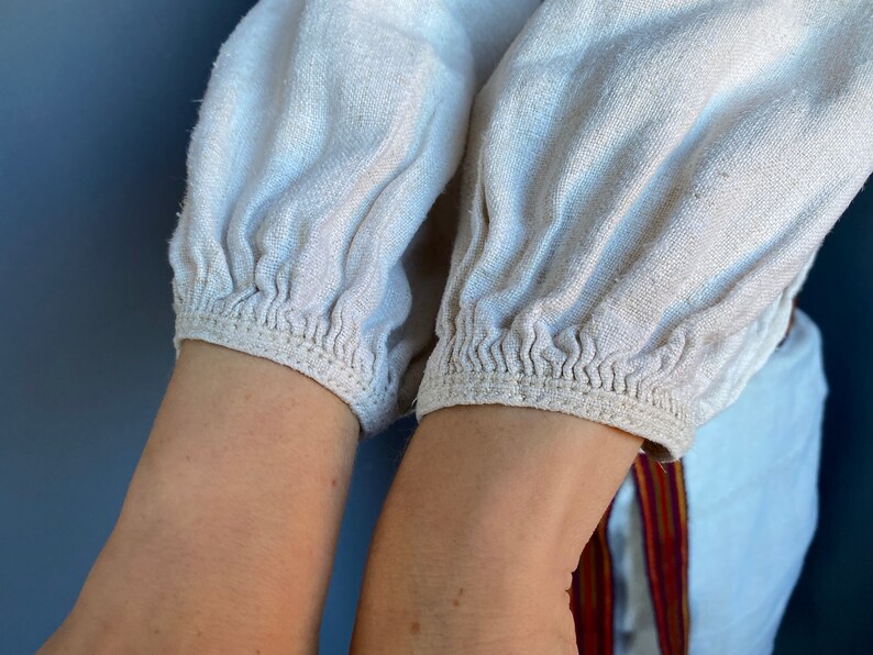 May include: A long-sleeved shirt made of off-white, textured fabric featuring gathered, elasticised cuffs at the wrists. The sleeves have a loose, voluminous fit, and a portion of a white garment with a vertical red and yellow striped trim is visible.