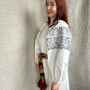 May include: A white embroidered linen dress with a colorful woven belt. The dress has a round neckline and long sleeves. The embroidery is in a floral pattern with blue, green, and white thread.