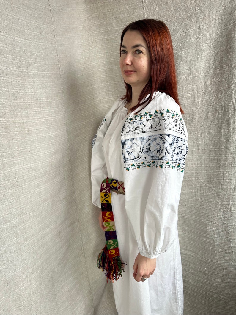 May include: A white embroidered linen dress with a colorful woven belt. The dress has a round neckline and long sleeves. The embroidery is in a floral pattern with blue, green, and white thread.