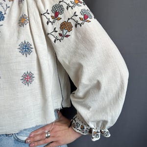 May include: White embroidered blouse with a floral pattern in shades of blue, red, orange, and black. The blouse has long sleeves with a gathered cuff and a loose fit.