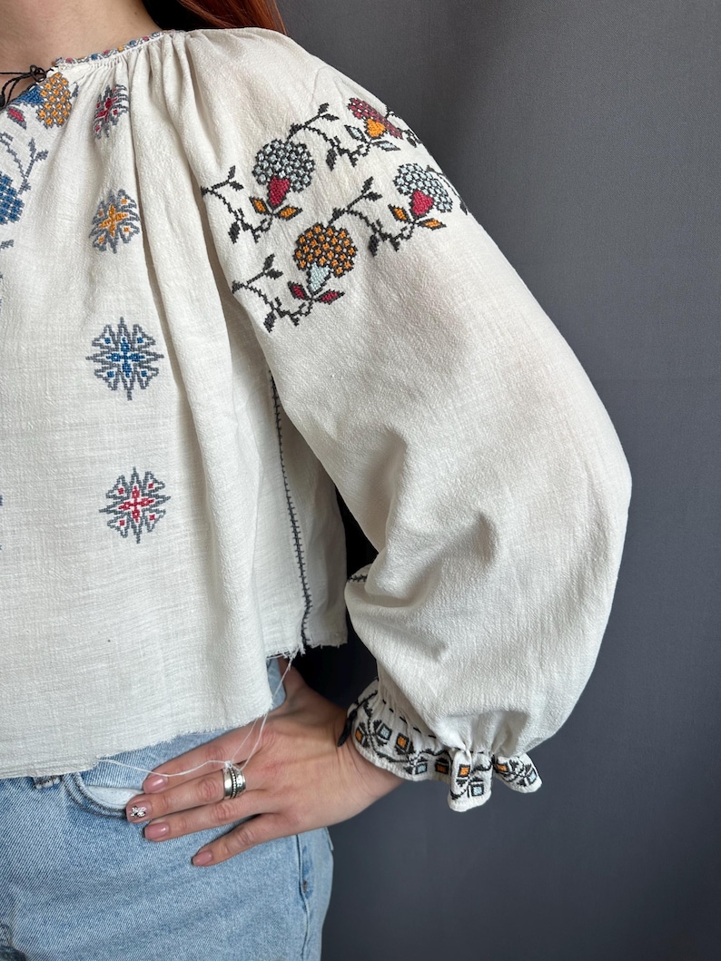 May include: White embroidered blouse with a floral pattern in shades of blue, red, orange, and black. The blouse has long sleeves with a gathered cuff and a loose fit.