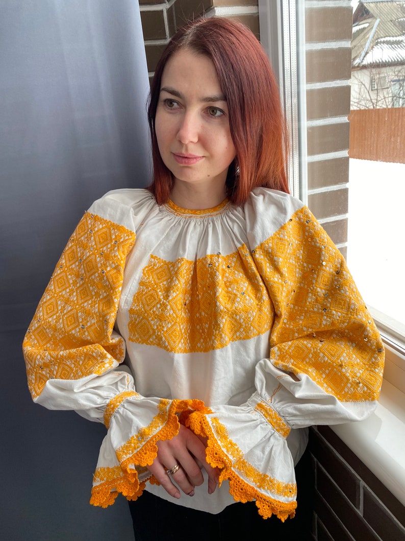 May include: A white embroidered blouse with yellow floral embroidery and crochet trim on the sleeves.