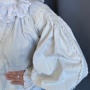 K&ouml;nnte beinhalten: Wei&szlig;e Leinenbluse mit R&uuml;schenkragen und langen &Auml;rmeln. Die &Auml;rmel haben einen gerafften Puff an der Schulter und sind mit Spitze besetzt.