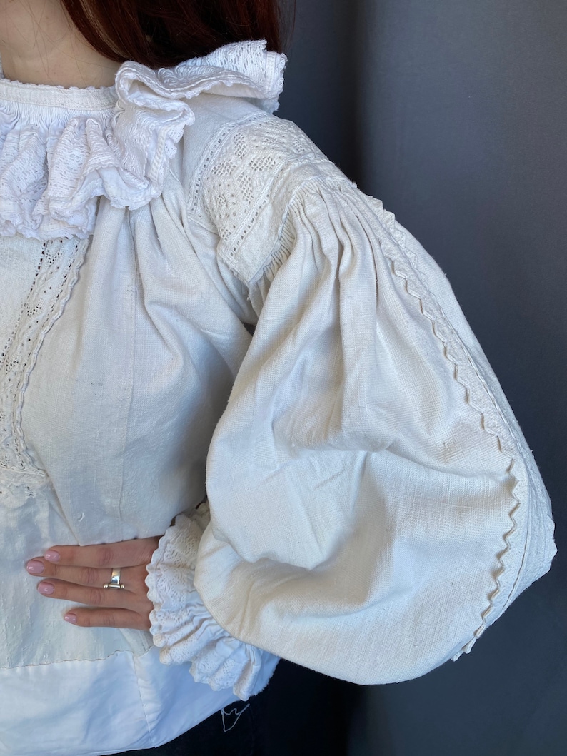 K&ouml;nnte beinhalten: Wei&szlig;e Leinenbluse mit R&uuml;schenkragen und langen &Auml;rmeln. Die &Auml;rmel haben einen gerafften Puff an der Schulter und sind mit Spitze besetzt.