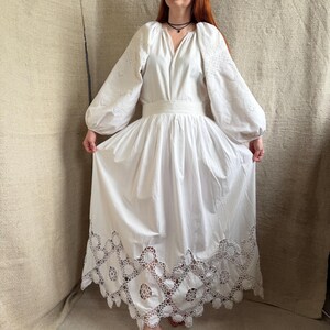 May include: A white dress with long, puffy sleeves and a V-neckline. The dress features intricate cutwork on the sleeves and a detailed, scalloped hemline. The waist is cinched with a matching fabric belt, creating a flowing, elegant silhouette.