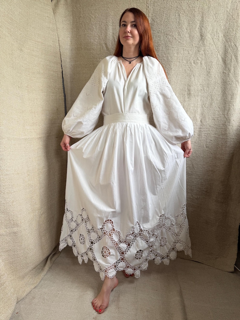 May include: A white dress with long, puffy sleeves and a V-neckline. The dress features intricate cutwork on the sleeves and a detailed, scalloped hemline. The waist is cinched with a matching fabric belt, creating a flowing, elegant silhouette.