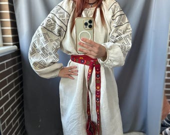 May include: A cream-colored linen dress with long sleeves featuring black geometric embroidery on the sleeves. The dress is cinched at the waist with a red embroidered belt with tassels. The dress is knee-length.