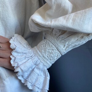 K&ouml;nnte beinhalten: Wei&szlig;e Leinenbluse mit einem ger&uuml;schten, bestickten &Auml;rmelbund. Der &Auml;rmelbund ist mit zarten Spitzendetails und einem gerafften R&uuml;schen verziert.