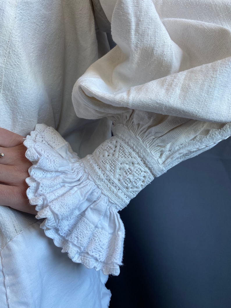 K&ouml;nnte beinhalten: Wei&szlig;e Leinenbluse mit einem ger&uuml;schten, bestickten &Auml;rmelbund. Der &Auml;rmelbund ist mit zarten Spitzendetails und einem gerafften R&uuml;schen verziert.