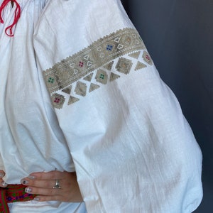 May include: White linen blouse with embroidered details in silver and colorful thread. The embroidery features a geometric pattern with squares and triangles.