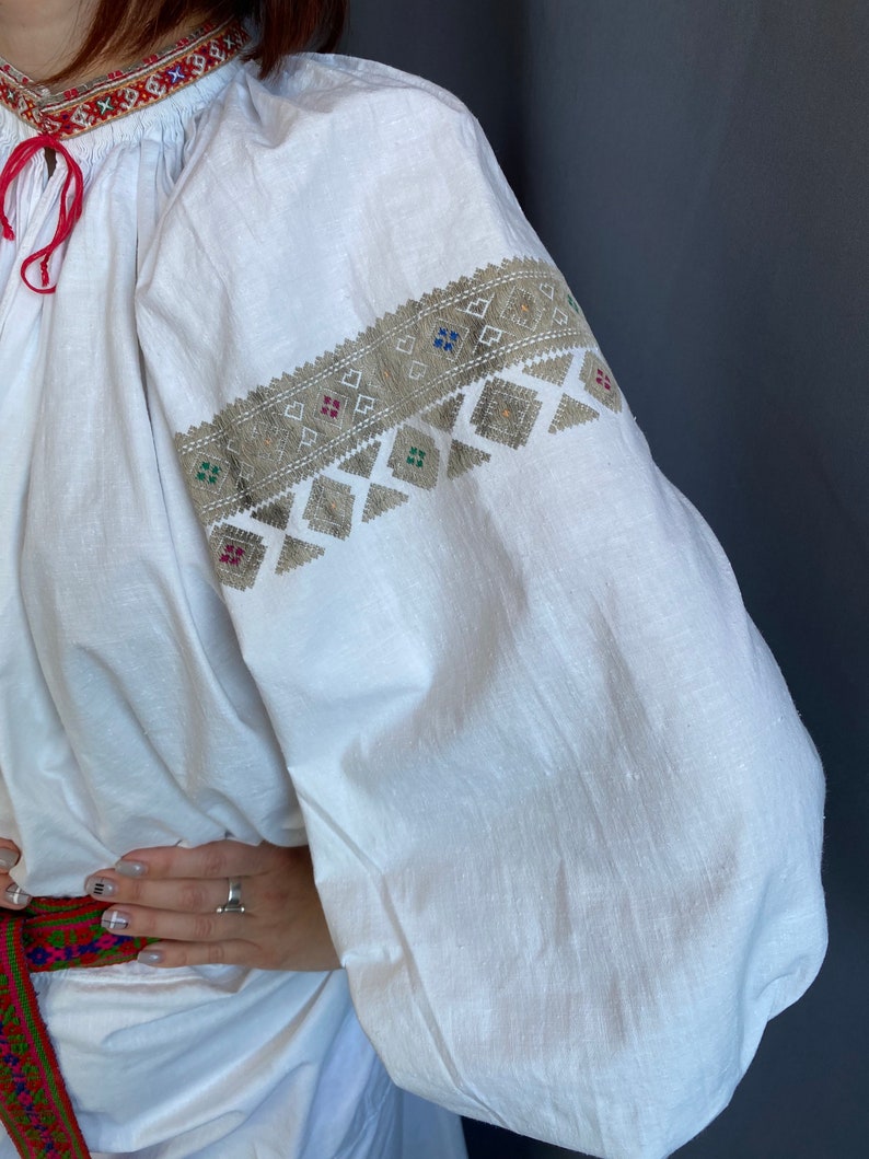 May include: White linen blouse with embroidered details in silver and colorful thread. The embroidery features a geometric pattern with squares and triangles.