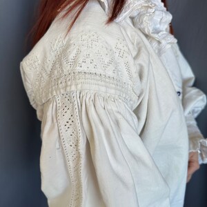 Op de afbeelding: Een wit linnen blouse met delicate kantdetails op de schouders en mouwen. De blouse heeft een gerimpelde kraag en een geplooide boord.