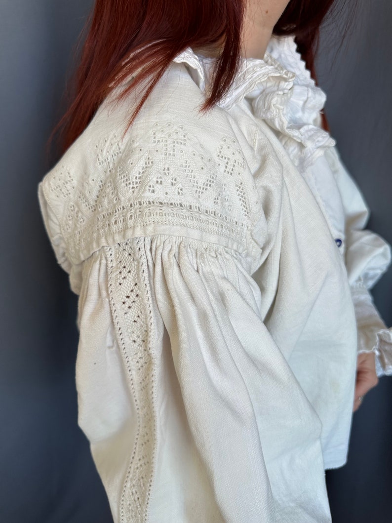Op de afbeelding: Een wit linnen blouse met delicate kantdetails op de schouders en mouwen. De blouse heeft een gerimpelde kraag en een geplooide boord.