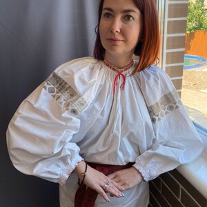 May include: A white embroidered blouse with long sleeves and a red ribbon tied at the neck. The blouse is worn with a red and blue embroidered belt.