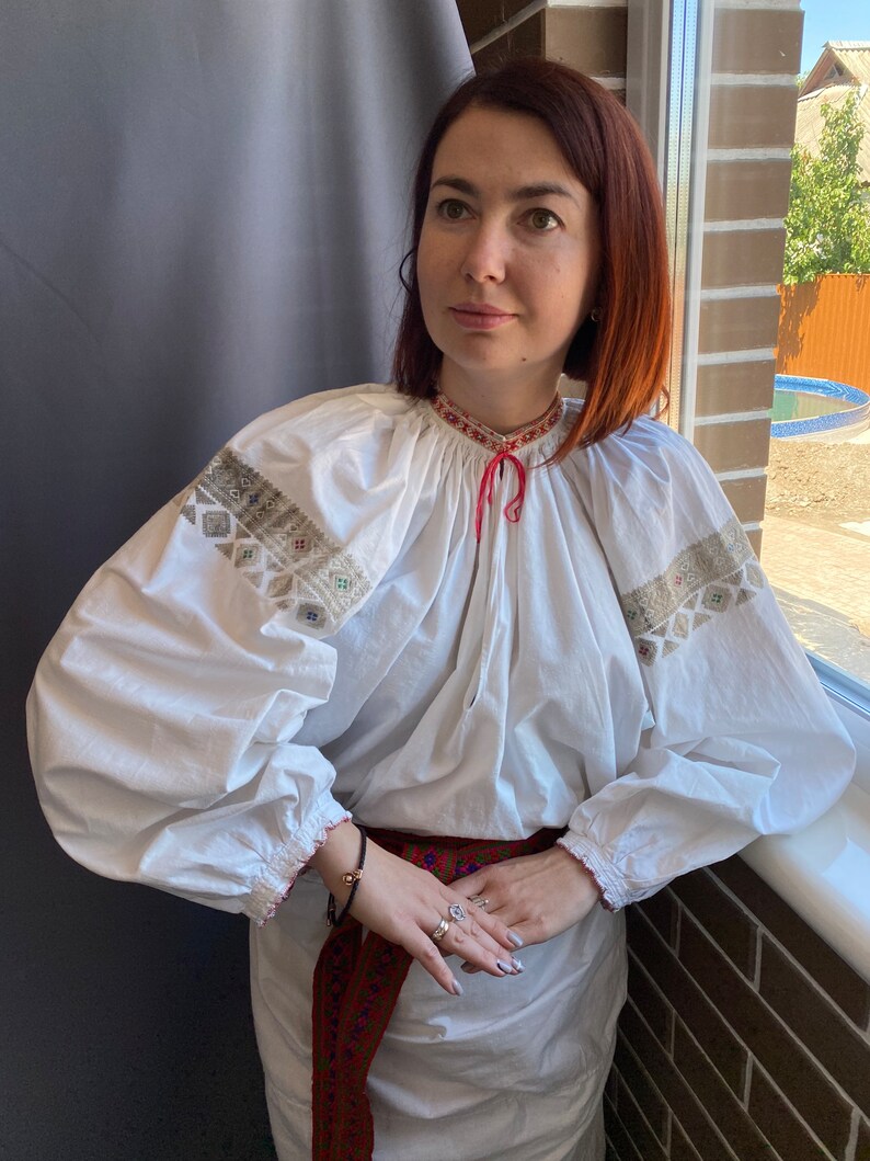 May include: A white embroidered blouse with long sleeves and a red ribbon tied at the neck. The blouse is worn with a red and blue embroidered belt.