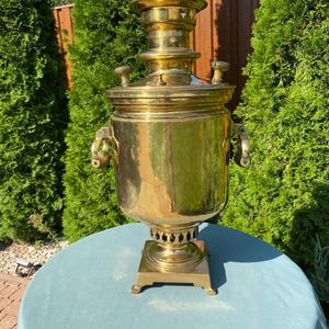 May include: A traditional brass samovar with a round body, a square base, and a tall spout. The samovar has two handles and a lid with a knob. It is sitting on a light blue tablecloth.