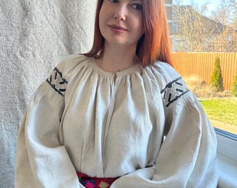 May include: A cream-colored linen dress with long, voluminous sleeves and black embroidered details on the shoulders. The dress is cinched at the waist with a colorful, patterned belt. The style is reminiscent of traditional Eastern European folk attire.